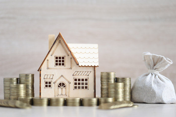 A wooden model house surrounded by stacks of coins and a small fabric money bag, representing real estate investment and financial growth.